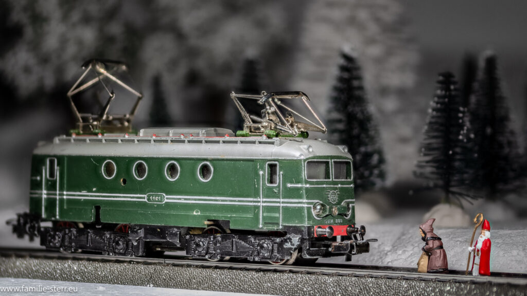 Nikolaus und Knecht Ruprecht stehen im Gleis und blockieren eine Lokomotive, die durch den winterlichen Wald fährt (Modellbahn)